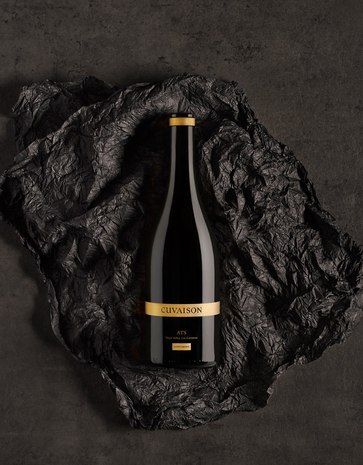 A dark glass bottle labeled Cuvaison Legacy Tasting Kit with gold accents sits on crumpled black textured paper against a dark stone background.