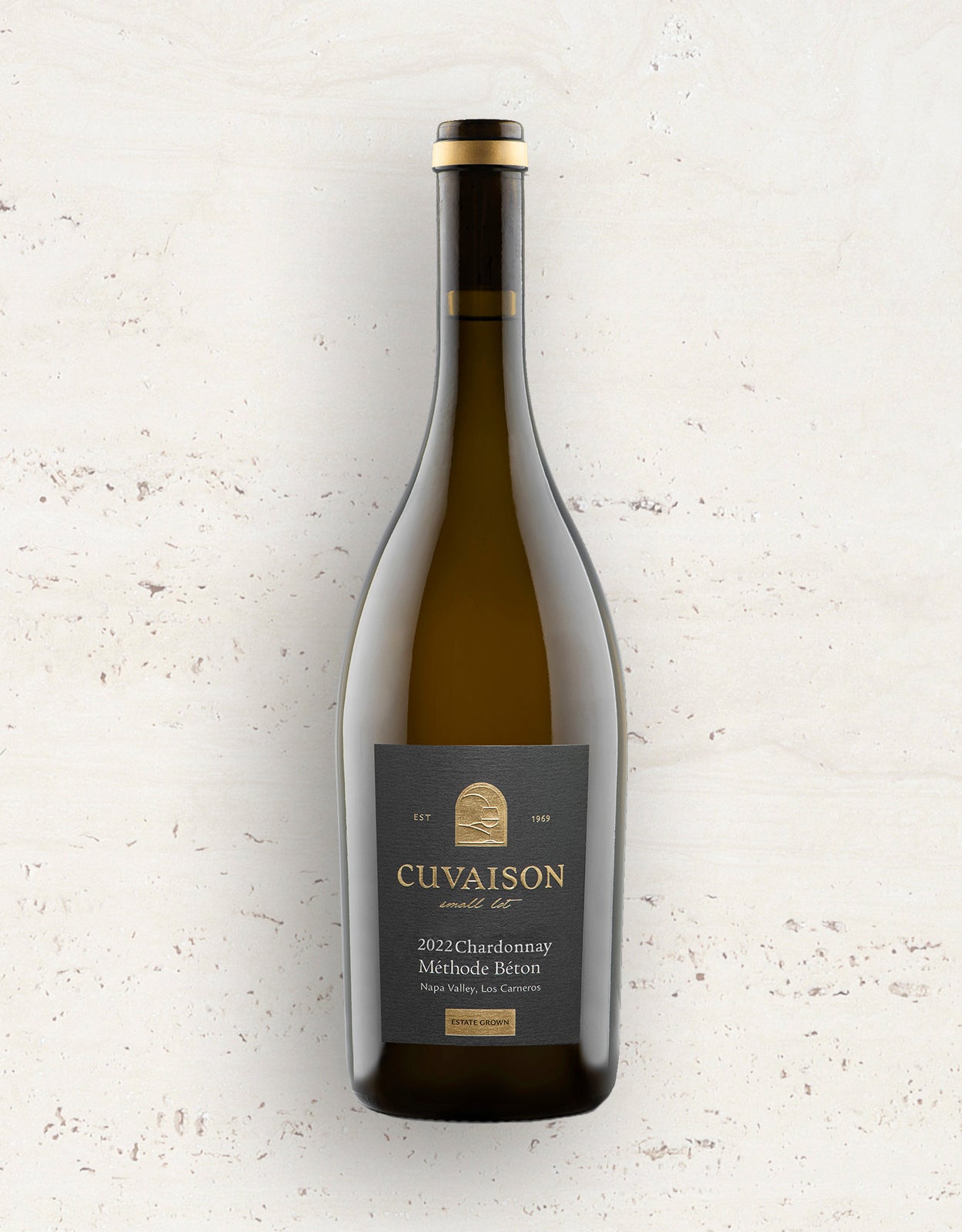 A bottle of 2022 Cuvaison Chardonnay, Méthode Béton stands upright against a light stone background, featuring a dark label with gold and white text plus a gold emblem near the neck.