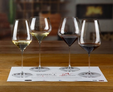 The Riedel Tasting Kit features four wine glasses with assorted white and red wines on a tasting mat atop a wooden surface, set against a softly blurred indoor background.