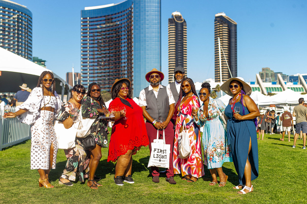 A group of people stand on grass at an outdoor event, posing for a photo. Tall modern buildings and white tents are visible in the background under a clear blue sky. Everyone is dressed in colorful, summery outfits.