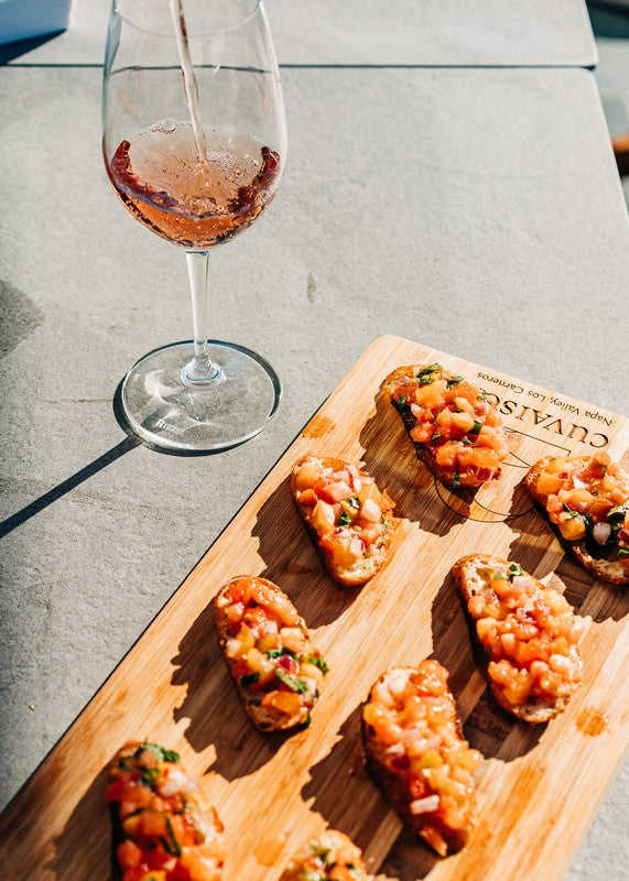 A glass of rosé wine is being poured beside a Cutting Board topped with several pieces of bruschetta with diced tomatoes and herbs, all placed on a gray table.