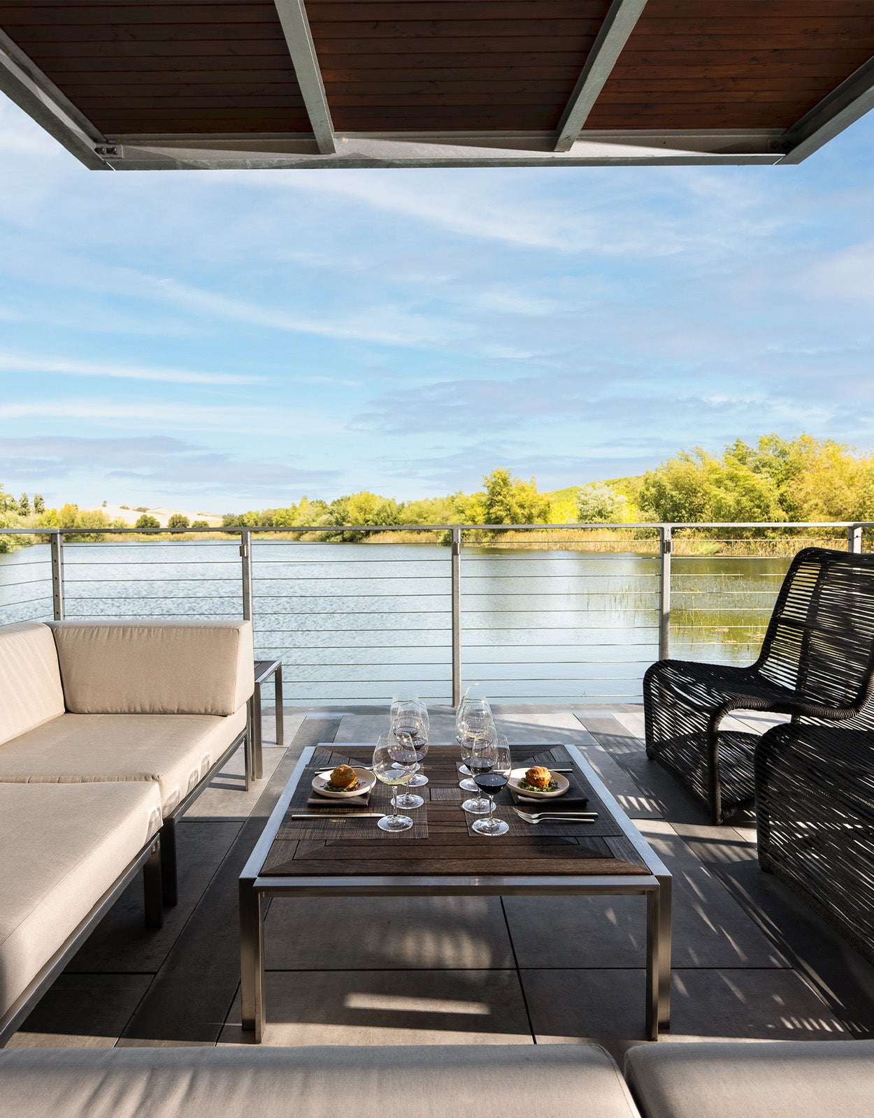 Enjoy The Boathouse Tasting Experience with comfortable beige cushioned seating and a black chair on a modern patio overlooking a tranquil river, surrounded by trees and blue sky, with wine glasses and small food bowls on the table.