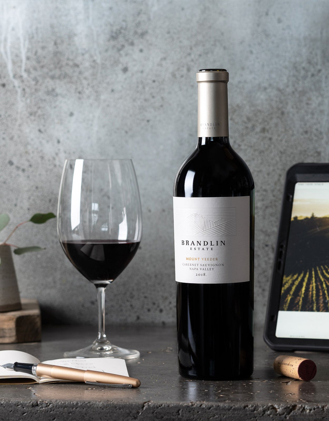 A Brandlin Estate Tasting Kit sits on a dark countertop, accompanied by a filled wine glass, a cork, a cheese knife, and a tablet showing a vineyard image, all set against a gray background.