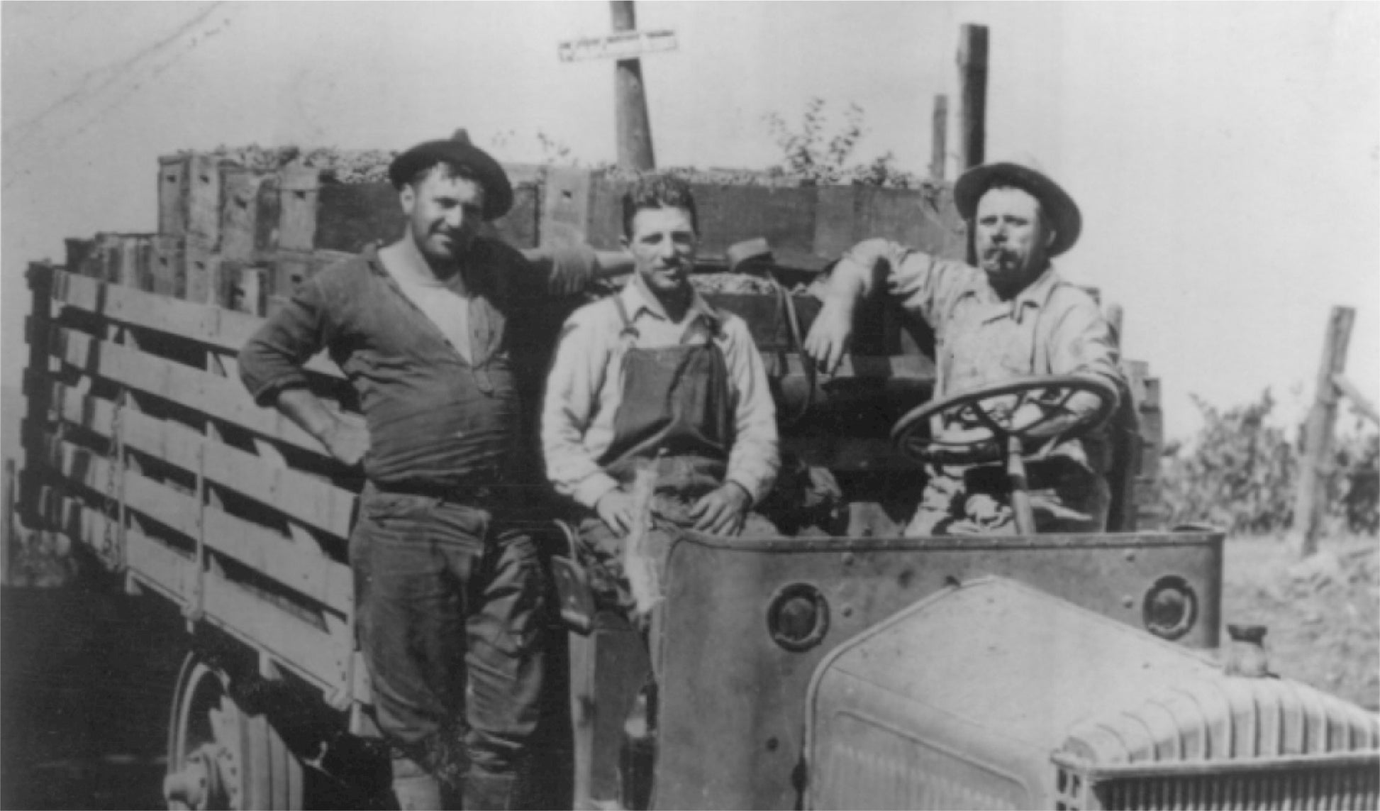 Three men wearing work clothes pose in and next to an old flatbed truck loaded with crates. The truck is parked outdoors, and there is a wooden post with a cross-shaped sign in the background.