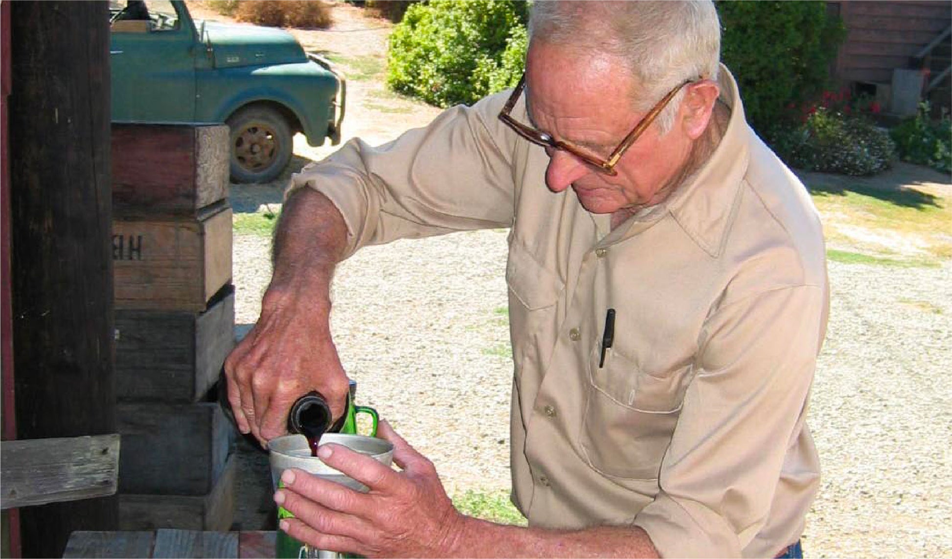 An older man with gray hair and glasses pours liquid from a dark bottle into a metal cup outdoors. He wears a beige shirt with a pen in the pocket. Wooden crates and a green truck are in the background.