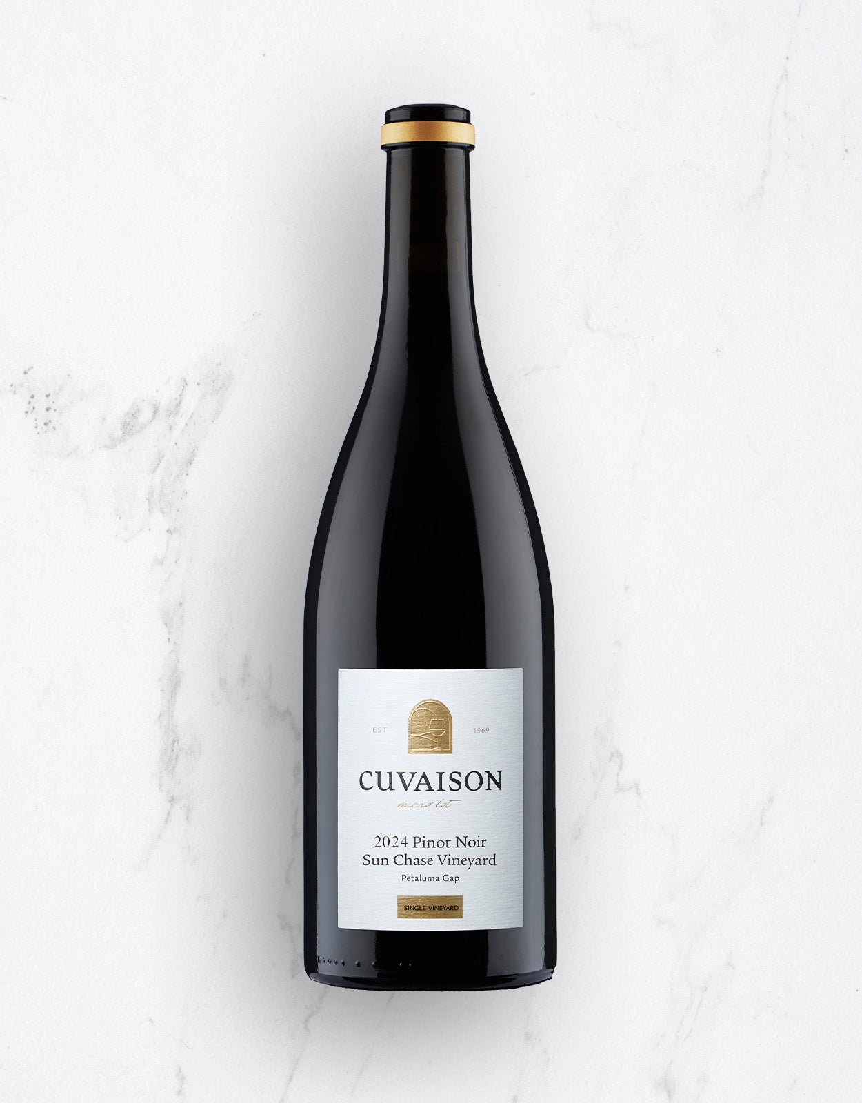 A bottle of 2024 Cuvaison Pinot Noir, Sun Chase Vineyard stands upright on a white marble surface. The label features white with gold and black accents.