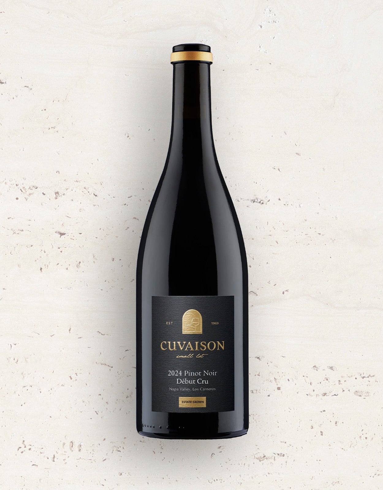 A bottle of 2024 Cuvaison Pinot Noir, Début Cru stands upright on a light, speckled surface. It features a black label with gold and white text and is topped with a gold cap.