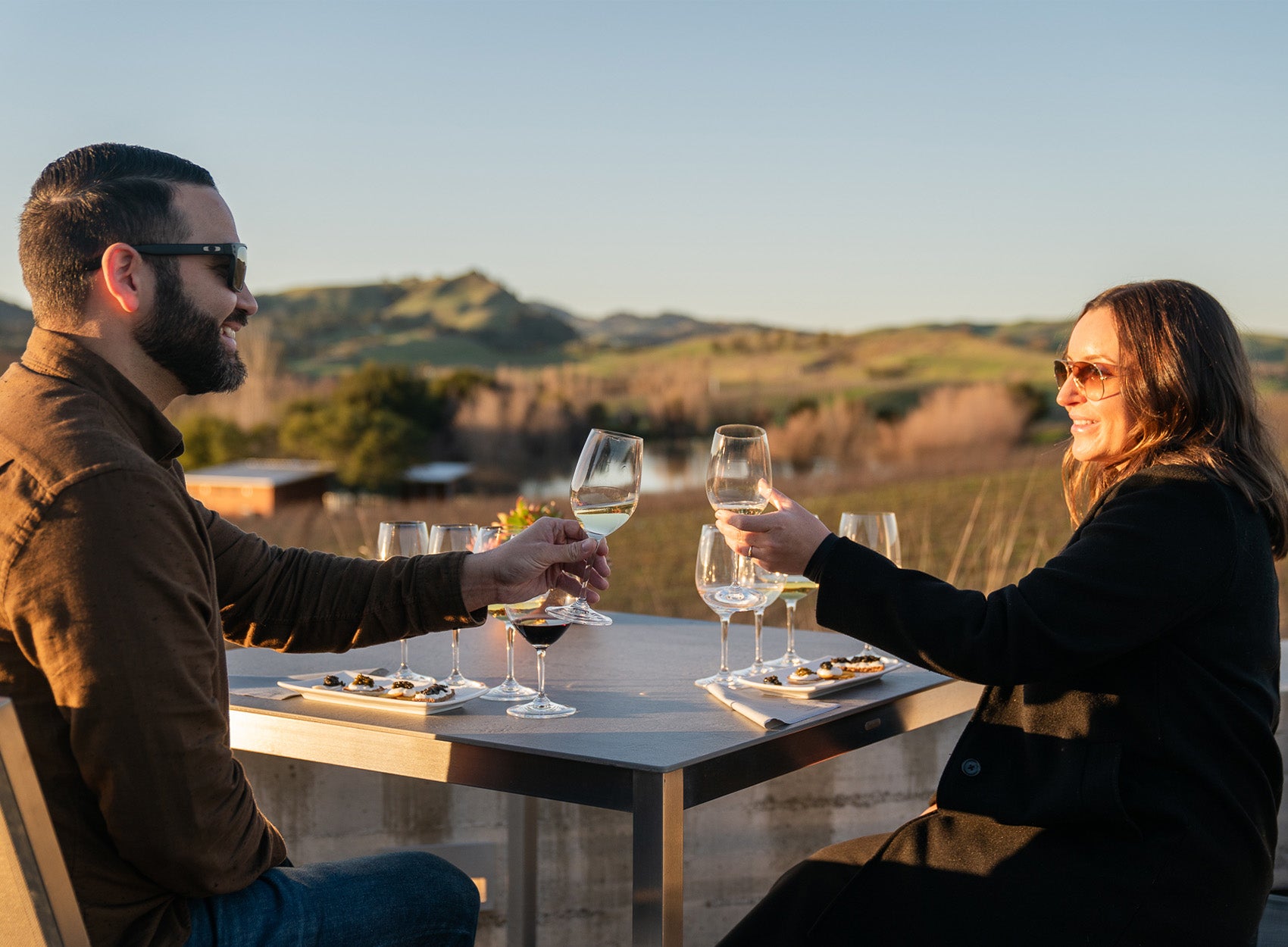 A man and woman sit at an outdoor table with wine glasses and small plates of food, toasting each other. They both wear sunglasses, and there are hills and greenery in the background.