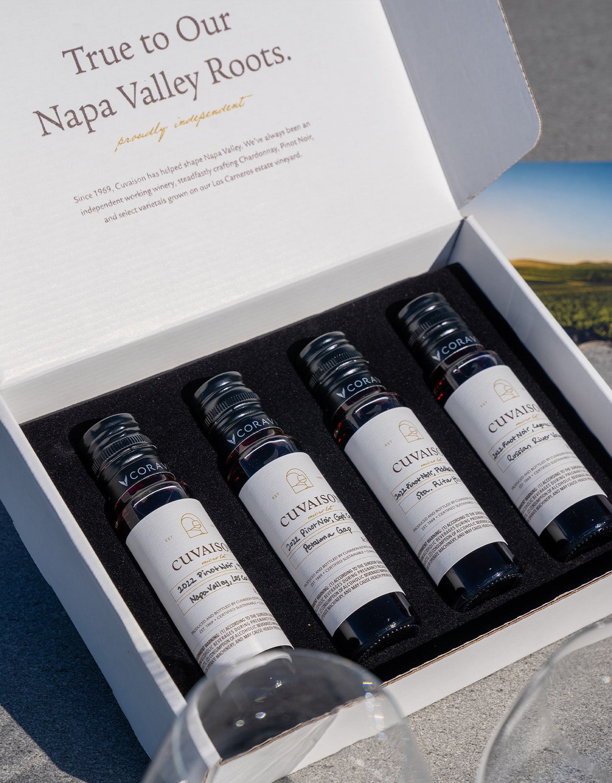 An open white box, labeled Cuvaison Micro Lot Tasting Kit, displays four different Napa Valley wine bottles. The lid features printed text about Napa Valley roots, and two empty wine glasses are partially visible.