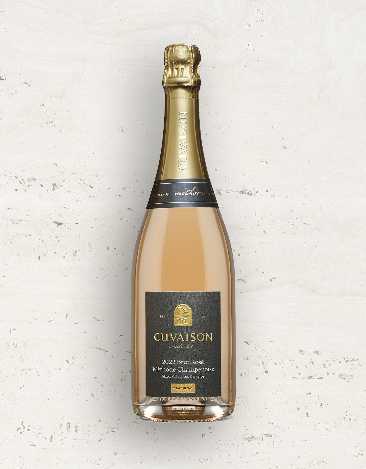 A bottle of 2022 Cuvaison Sparkling Brut Rosé with a gold foil top and black-and-gold label stands upright against a light textured background.