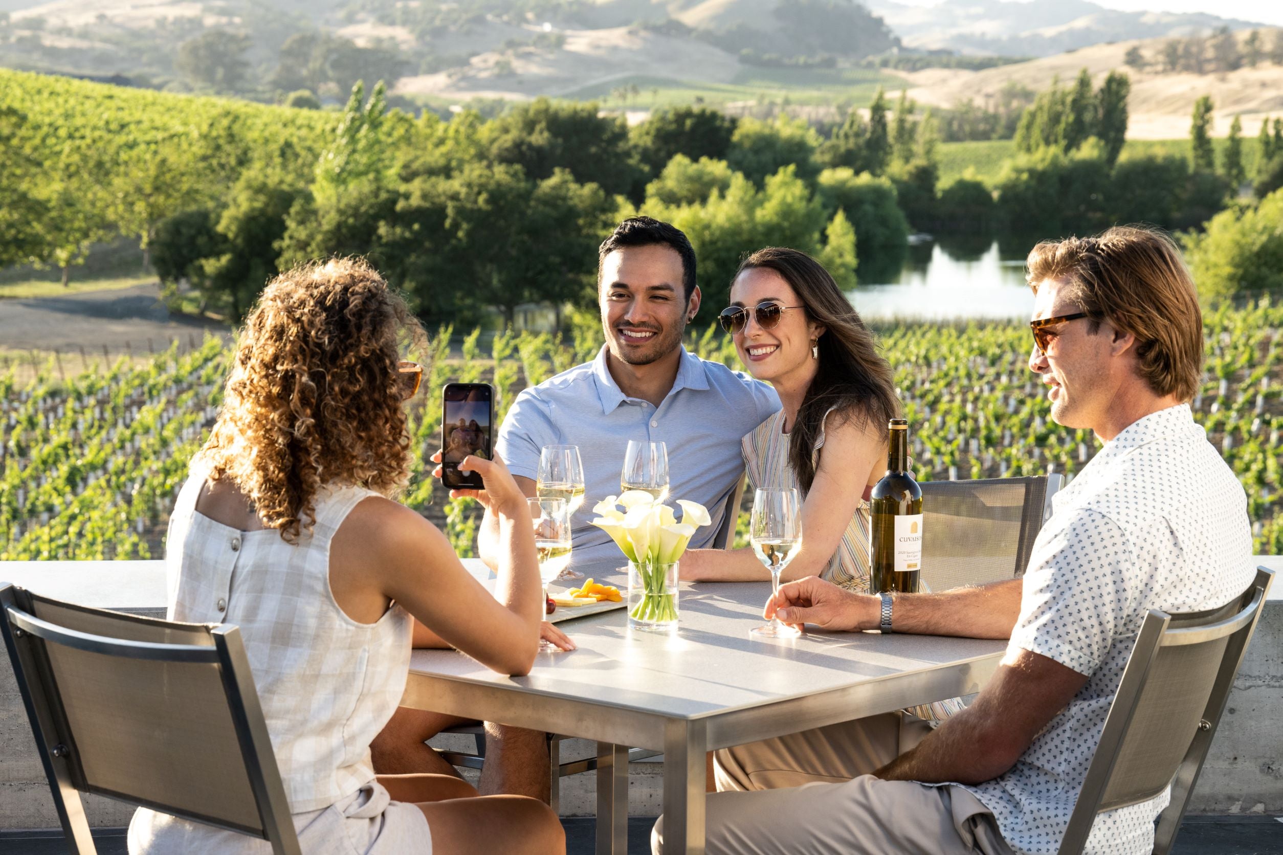Four adults sit at a patio table with wine glasses, a bottle of wine, and flowers, surrounded by vineyards and hills. One person takes a photo of the others, who are smiling at the camera.