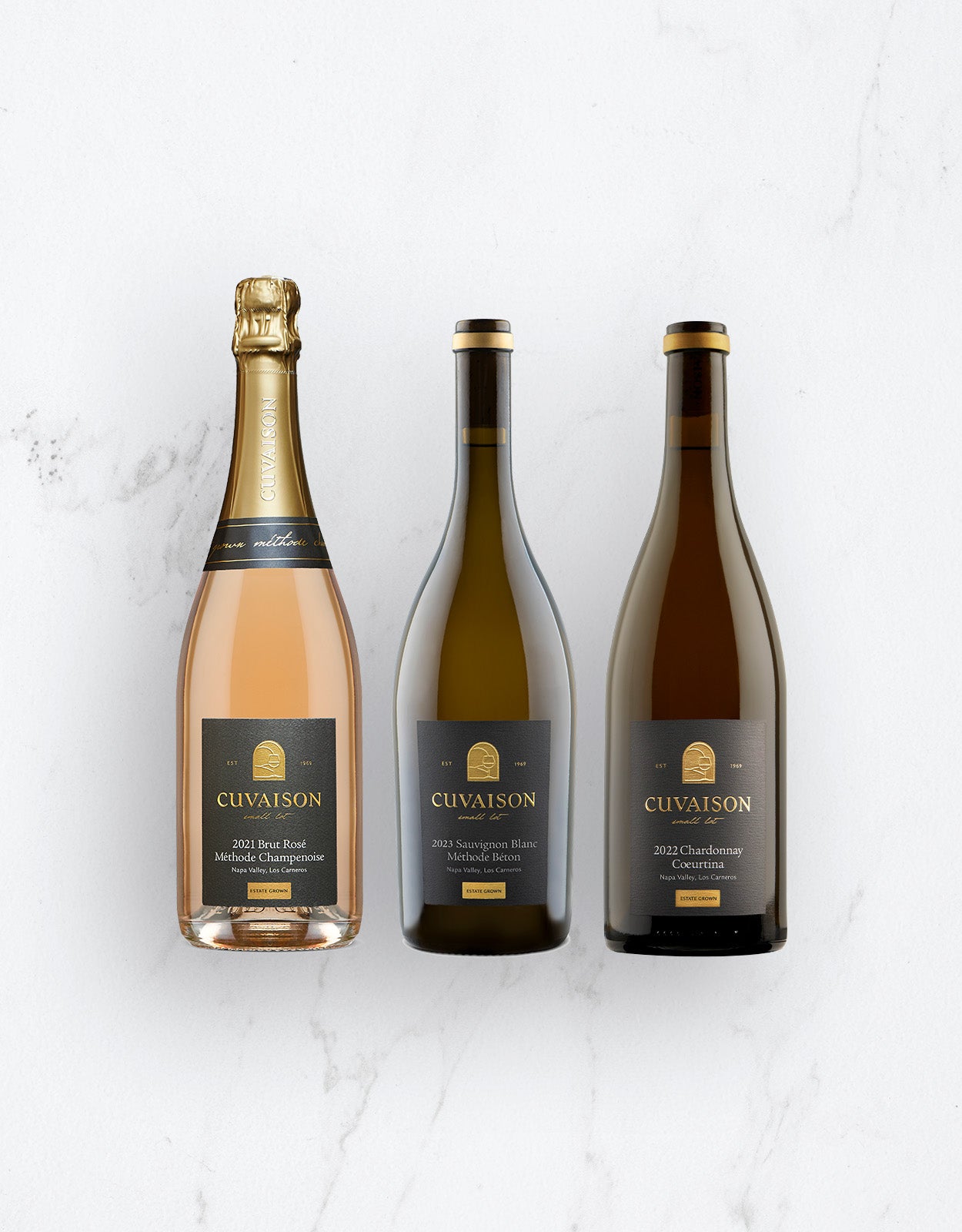 The Cuvaison Spring Table Trio is displayed side by side on a white surface: sparkling rosé on the left, Sauvignon Blanc in the middle, and Chardonnay on the right. Each bottle features a dark label with gold accents.
