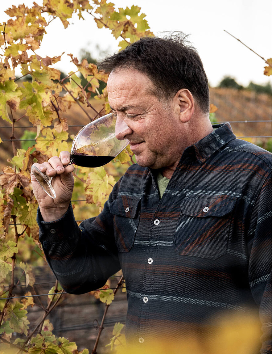 A man wearing a plaid shirt holds a glass of red wine near his nose, smelling it, while standing among grapevines with yellow leaves in a vineyard.