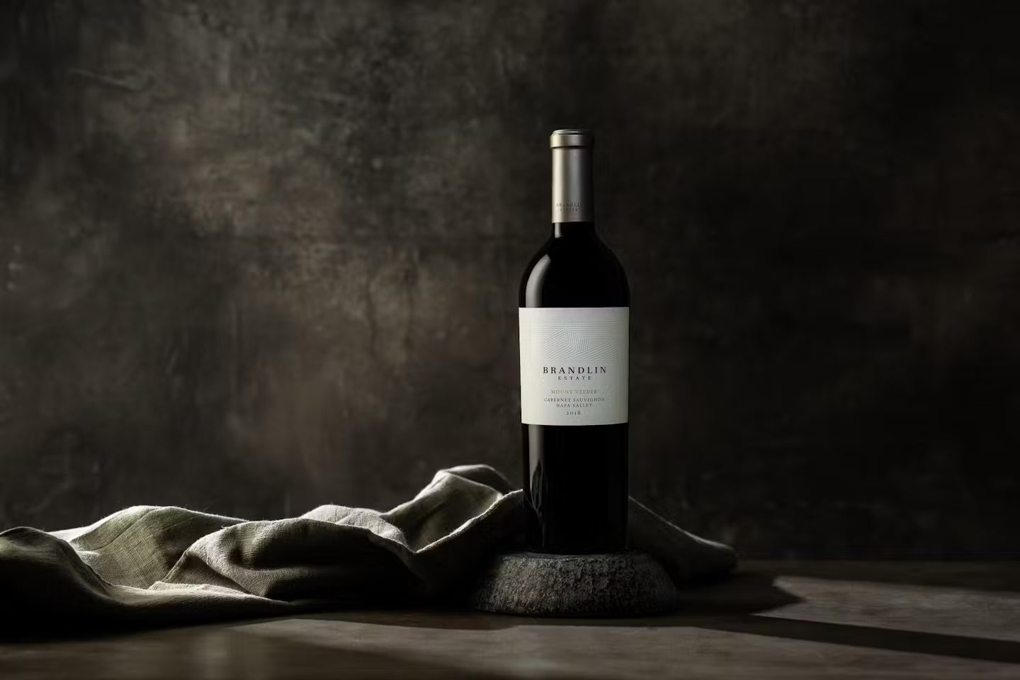 A bottle of Brandlin wine stands upright on a stone surface next to a folded cloth, set against a dark, textured background with dramatic lighting highlighting the bottle.