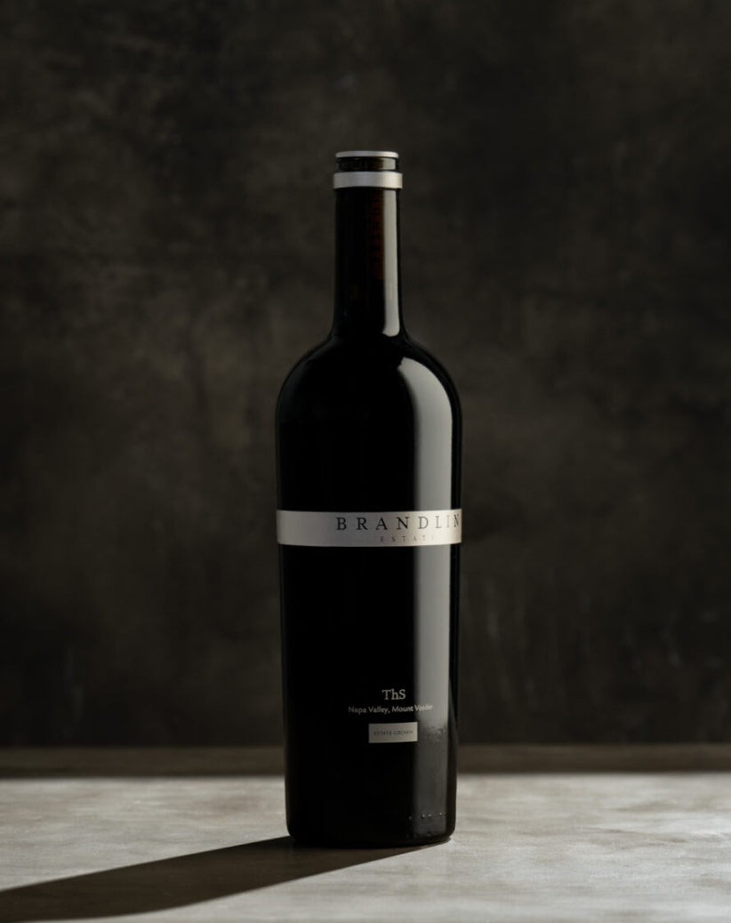 A dark glass wine bottle with a metallic label reading Brandlin stands upright on a stone surface against a dark, textured background. The lower label shows smaller text, including Napa Valley, Mount Veeder.