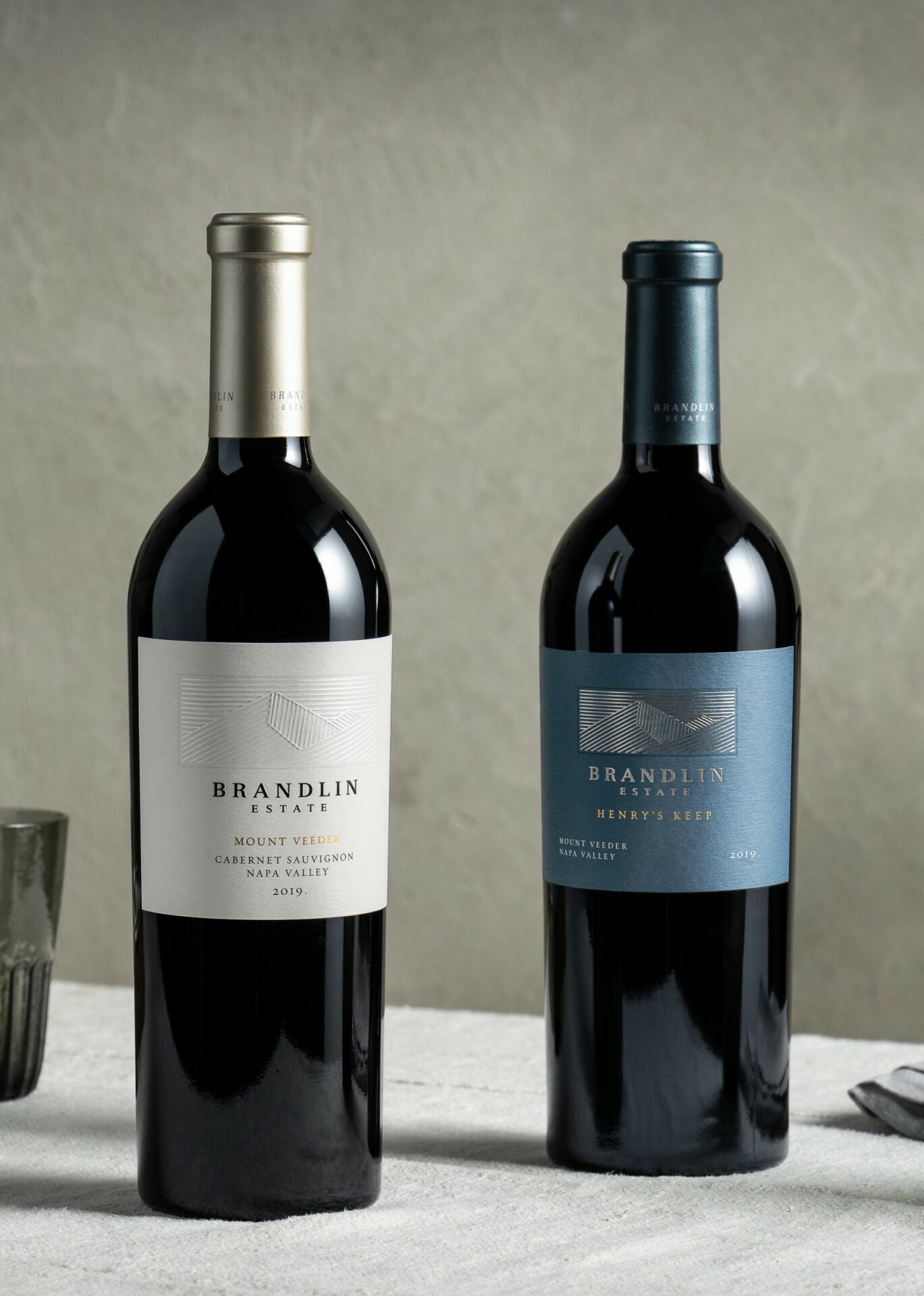 Two bottles of Brandlin Estate red wine stand side by side on a textured white surface, each with a different label and blue-gray background. A glass is partially visible on the left.