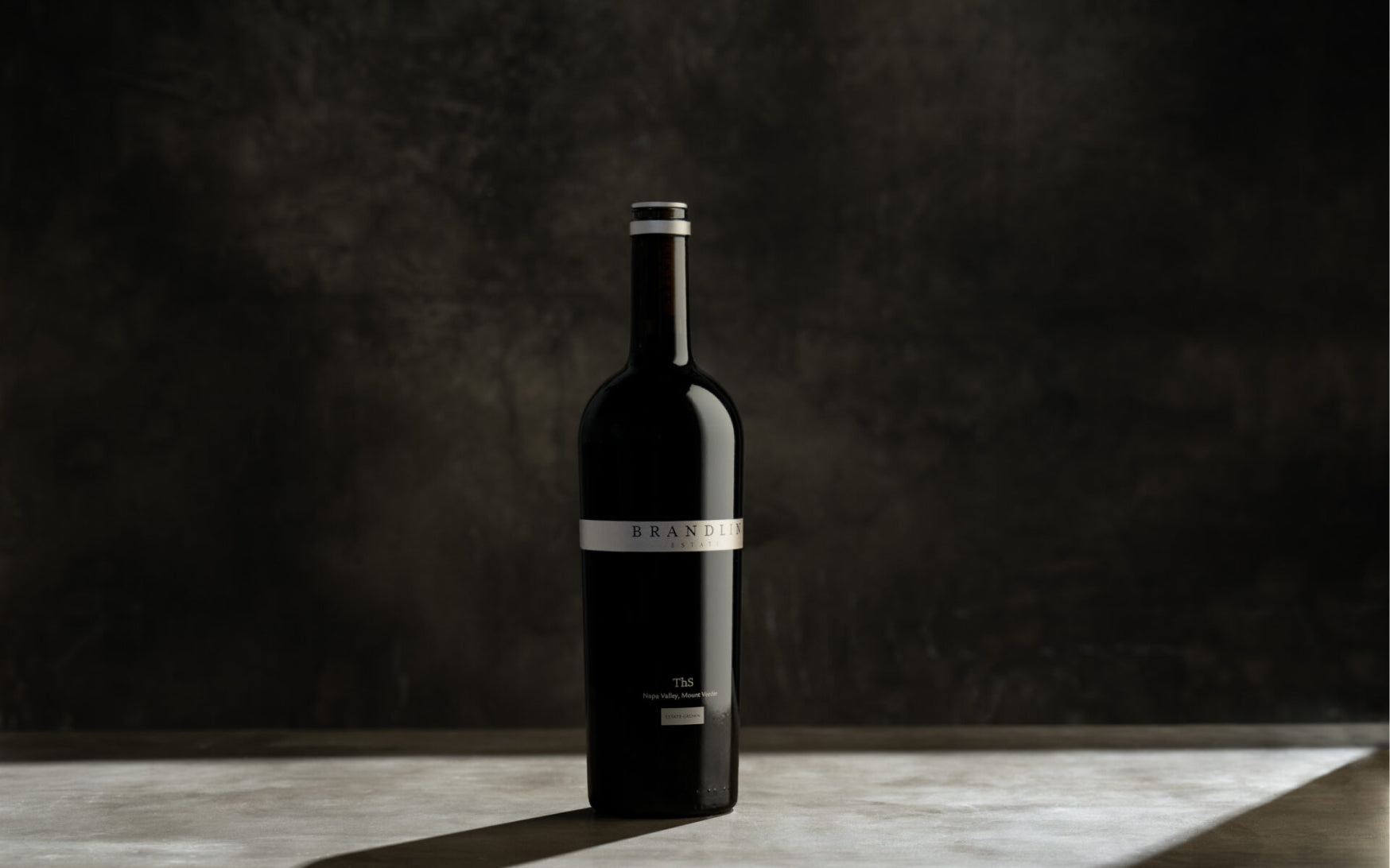 A single dark glass wine bottle labeled Brandlin 12 Bottle Club stands upright on a smooth surface with a dark, textured background.