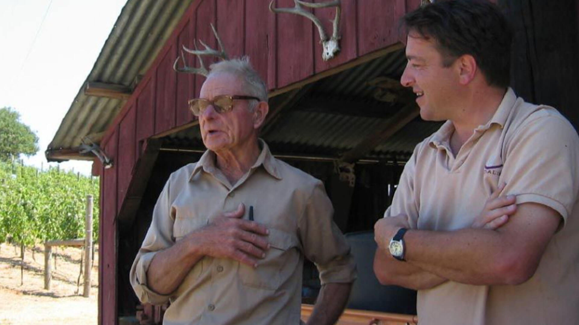Two men stand outside a red wooden building with antlers mounted above the door. One older man gestures with his hand on his chest while the younger man stands with arms crossed, both wearing light-colored shirts. A vineyard is in the background.