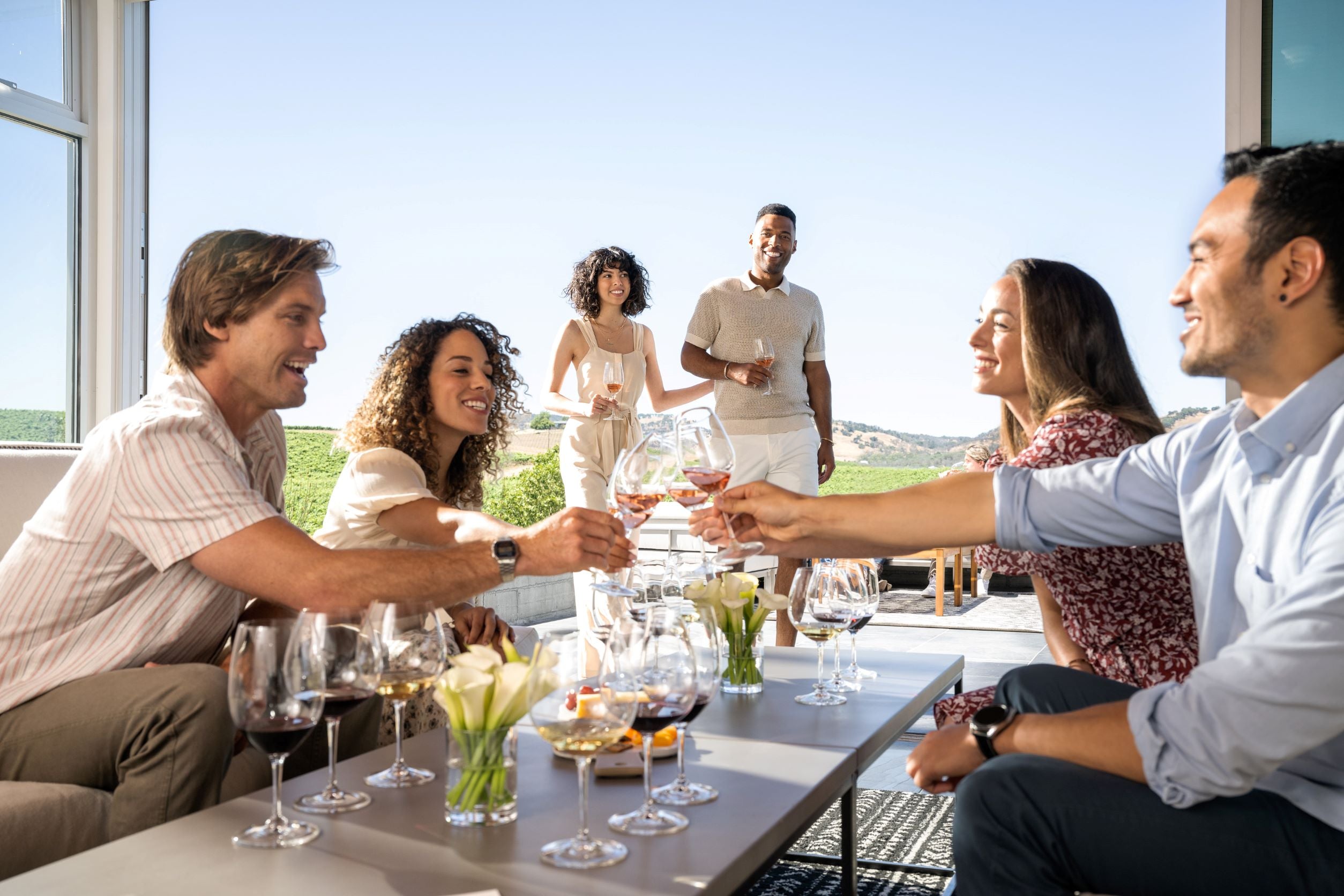 Six adults gather on a patio, clinking wine glasses around a table with more wine glasses and vases of white flowers. Two people stand in the background, while four sit and toast. The setting appears sunny and outdoors.