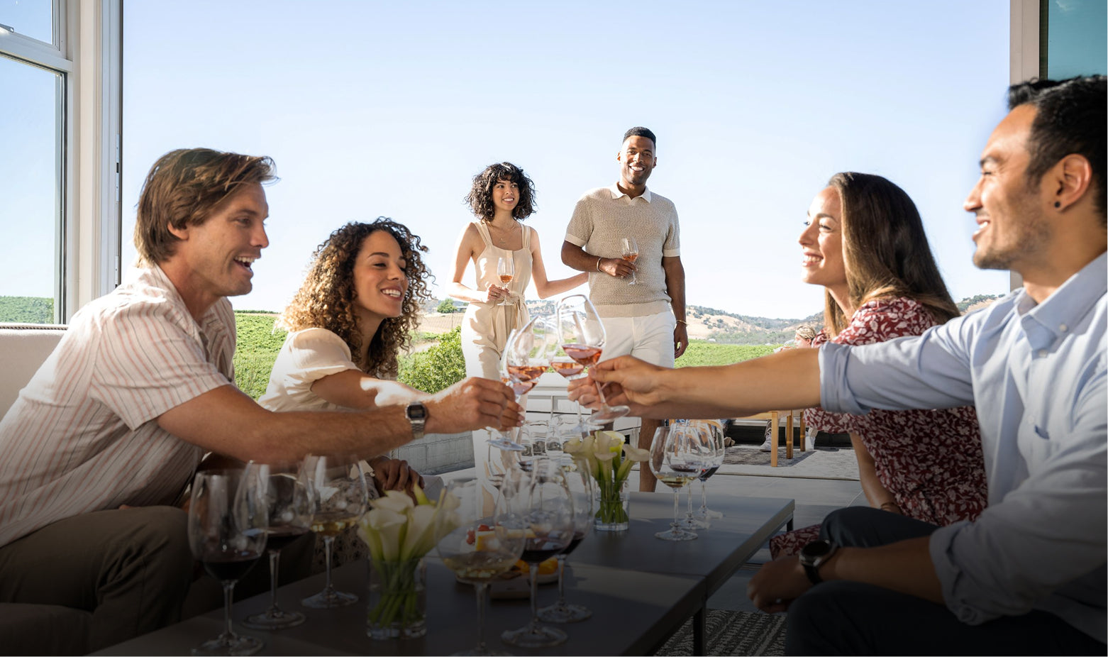 Six adults enjoy a wine tasting outdoors, clinking glasses and smiling. Two people stand in the background, while four sit at a table with wine glasses and flowers. Vineyards and hills are visible in the distance.