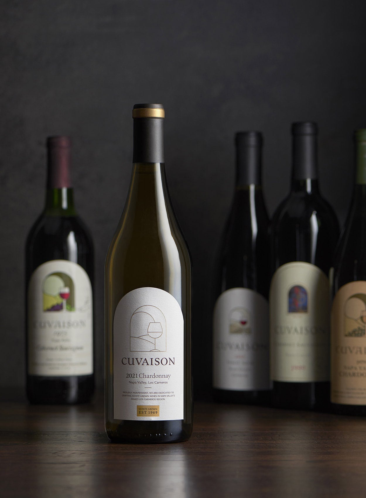 A bottle of CuvaIson 2021 Chardonnay is centered in the foreground, with four other assorted bottles of wine slightly out of focus in the background against a dark backdrop.