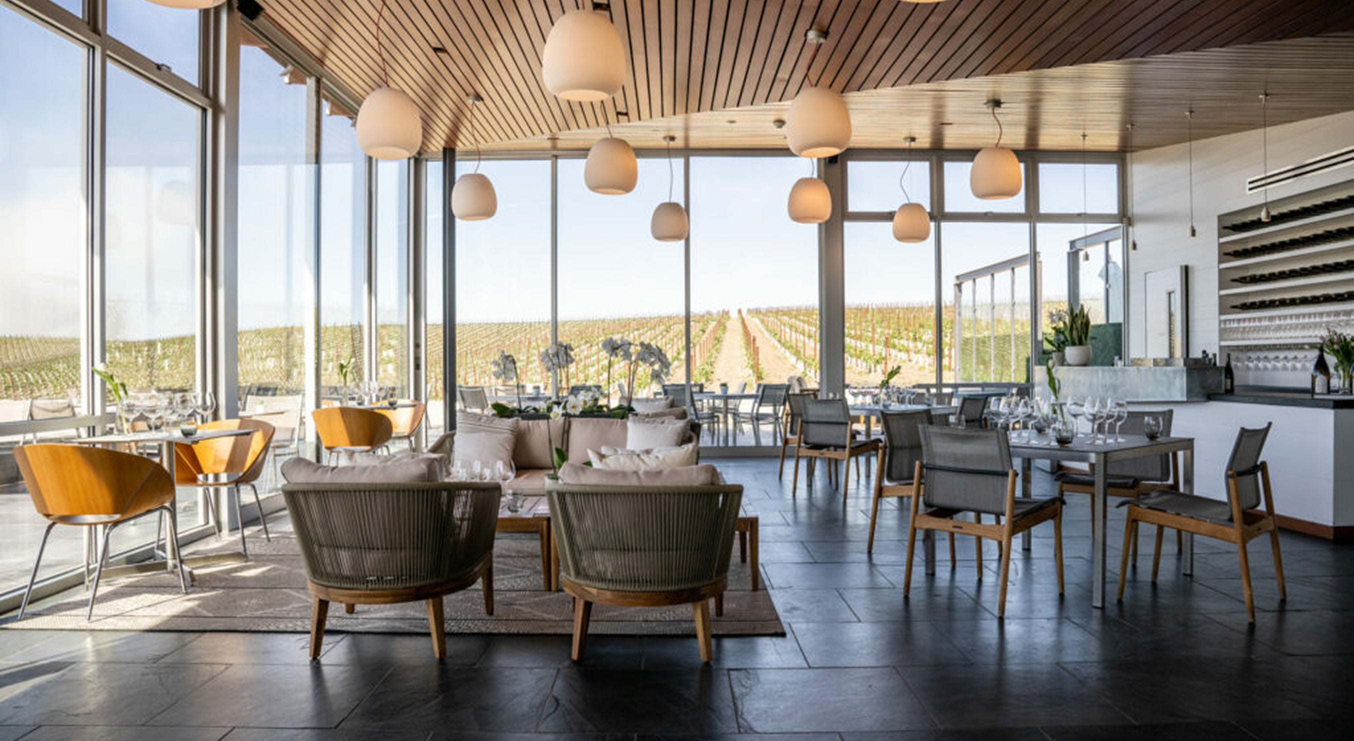 Modern restaurant interior with large floor-to-ceiling windows overlooking a vineyard, wooden ceiling, pendant lights, neutral-colored chairs and tables, and place settings ready for guests.