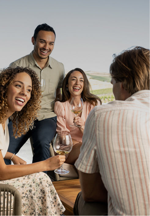 Four adults gather outdoors, smiling and holding wine glasses of 2021 Brandlin Estate Cabernet Sauvignon ThS. Against an open landscape with clear skies, they enjoy a relaxed social moment together.