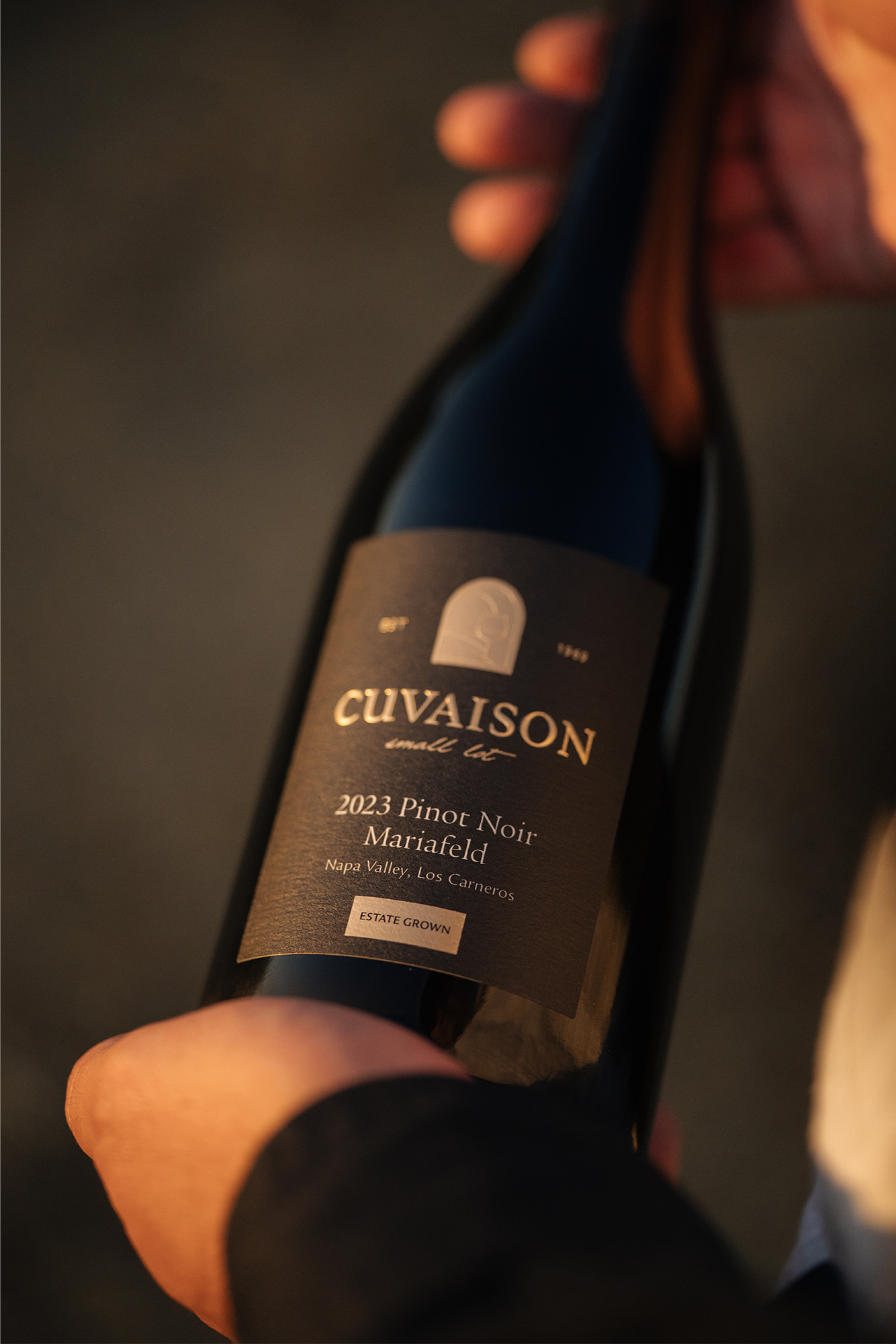 A person holds a bottle of Cuvaison 2023 Pinot Noir wine, labeled Mariafeld, Napa Valley, Los Carneros, Estate Grown. The label is dark and features gold and white text.