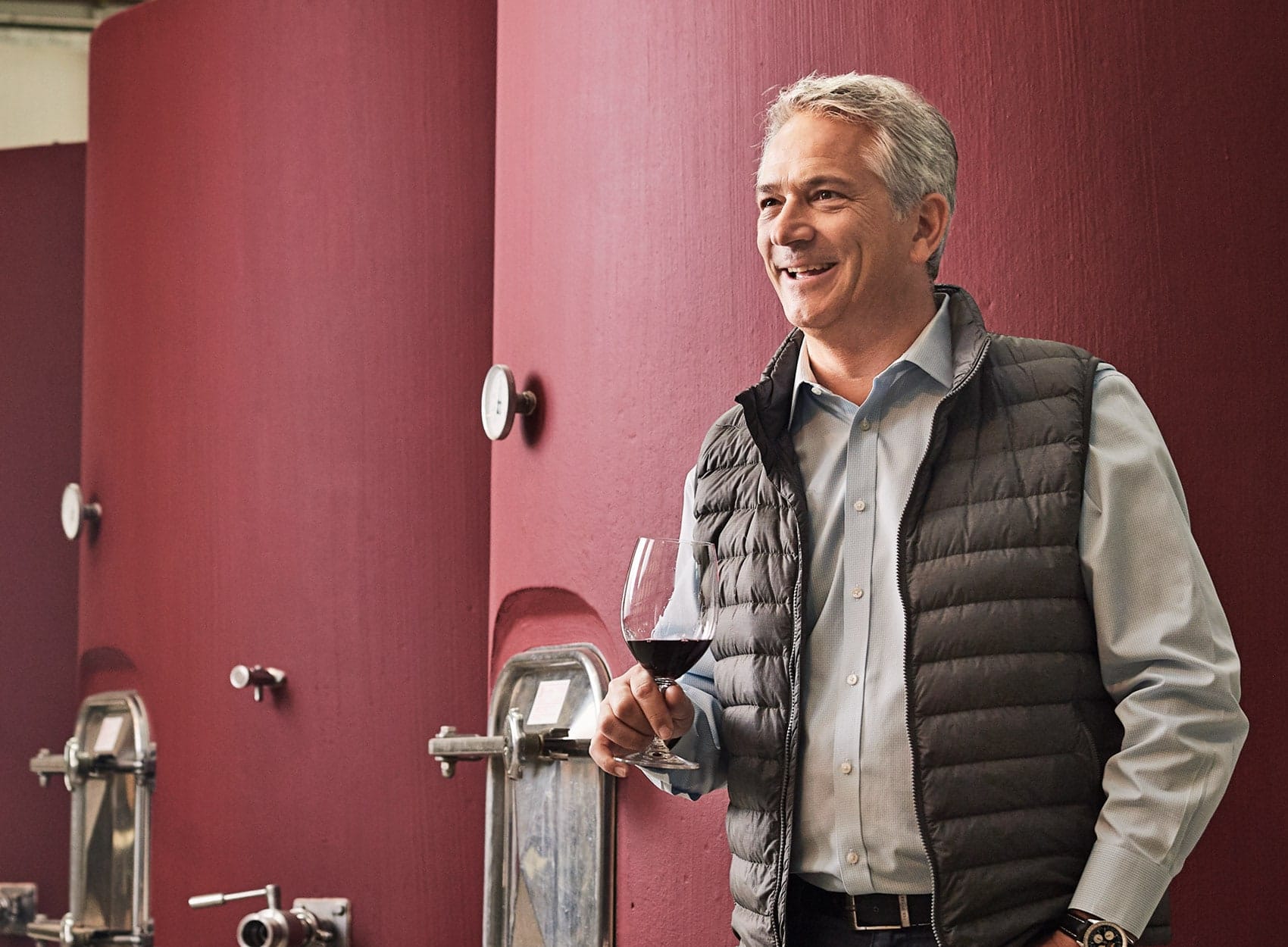A man with gray hair wearing a blue shirt and dark vest holds a glass of red wine and smiles while standing in front of large red wine fermentation tanks.