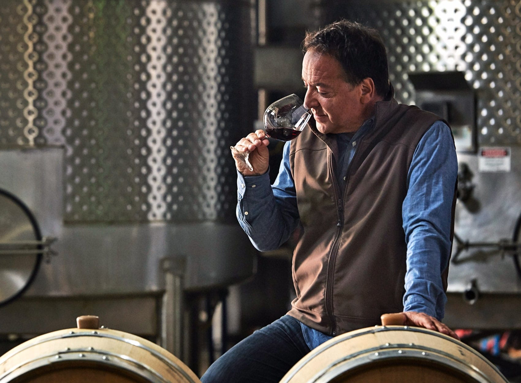 A man in a blue shirt and brown vest sits on a barrel in a winery, holding a glass of red wine to his nose. Large metal fermentation tanks are seen in the background.