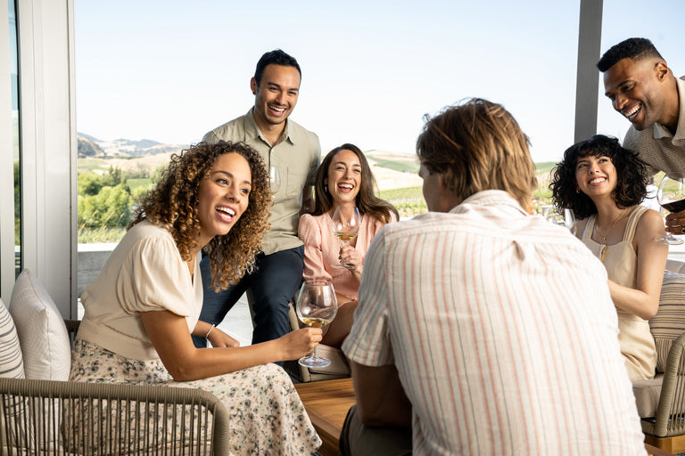 Six adults gather on a sunny patio with drinks, smiling and chatting around a table. Several enjoy glasses of 2018 Cuvaison Blanc de Blanc, Méthode Champenoise, with scenic landscape views in the background.
