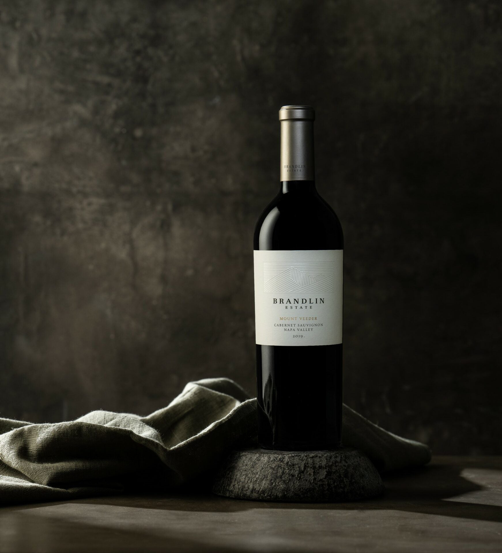 A bottle of Brandlin Estate Cabernet Sauvignon stands upright on a stone platform, with a folded gray cloth beside it against a dark, textured background.