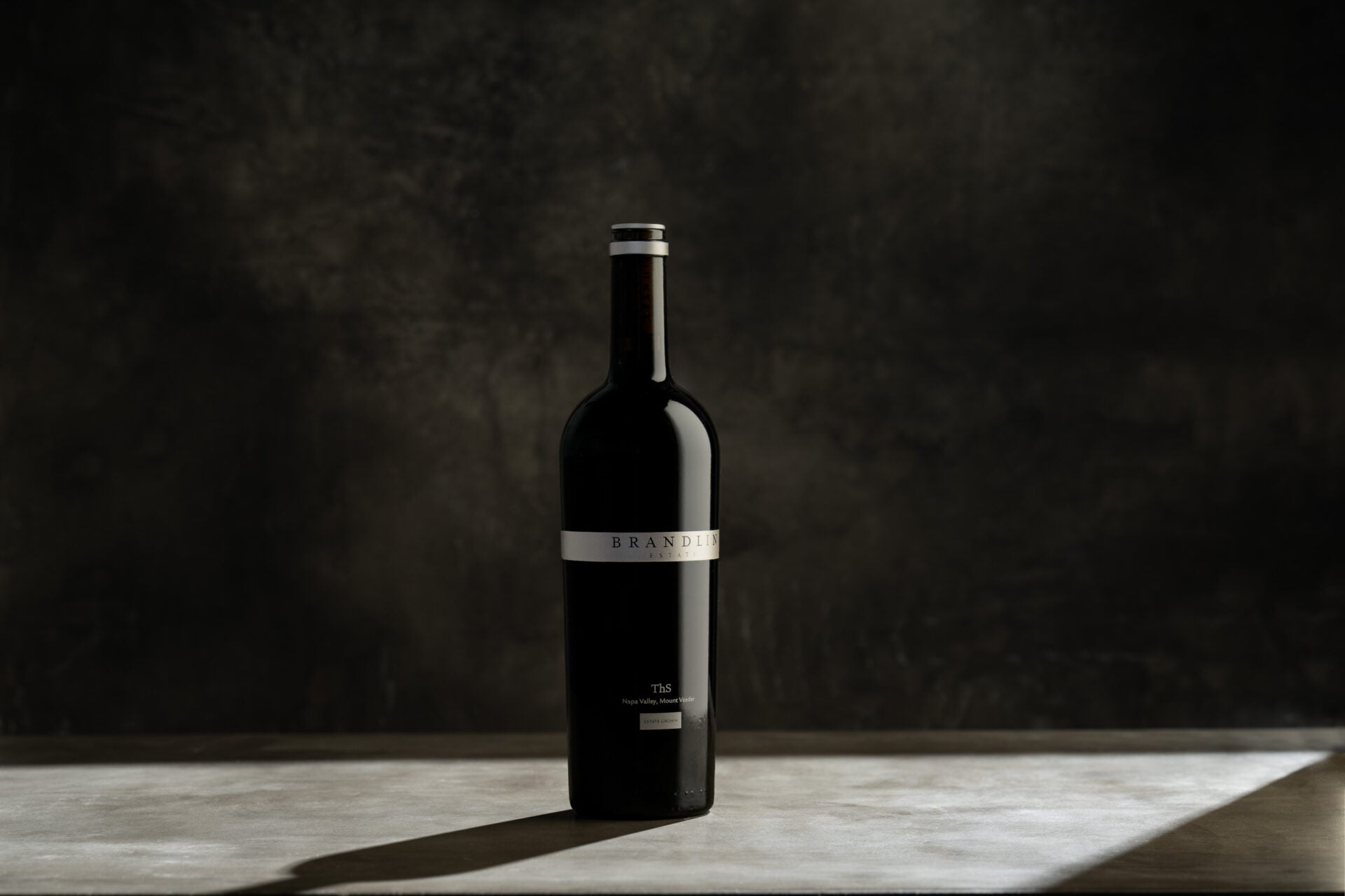 A dark glass wine bottle stands upright on a light surface against a dark, textured background. The label is minimal and partially visible, with BRANDIN readable in the center. Light creates a shadow beside the bottle.
