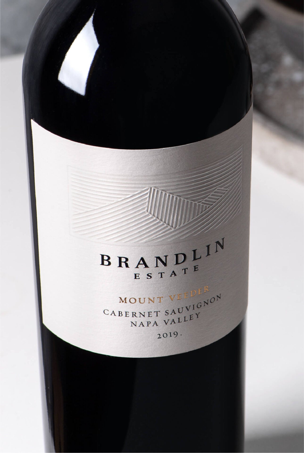 A close-up of a wine bottle labeled “Brandlin Estate Mount Veeder Cabernet Sauvignon Napa Valley 2019.” The label is off-white with embossed geometric lines and gold text accents.