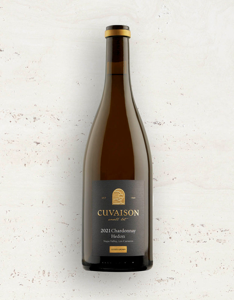 A bottle of 2021 Cuvaison Chardonnay, Hedon with a dark label and gold accents stands upright on a light, speckled surface.