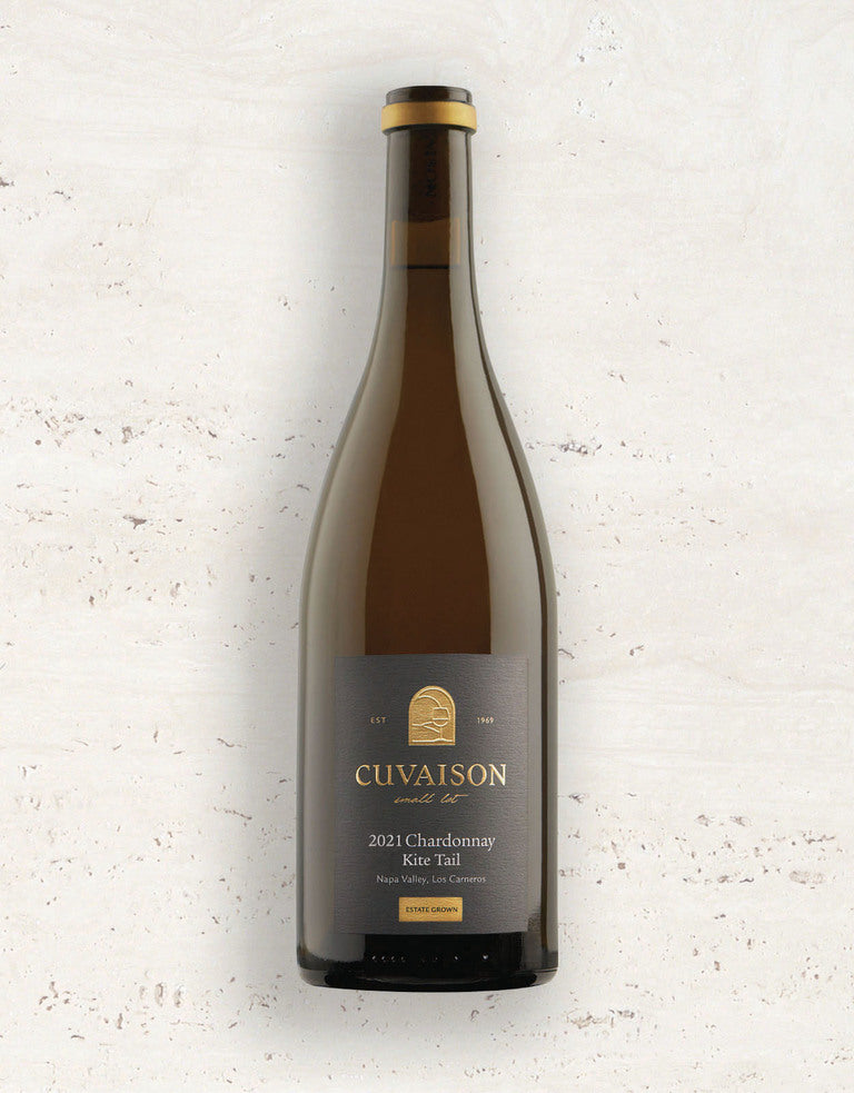 A bottle of 2021 Cuvaison Chardonnay, Kite Tail stands upright on a light textured surface with a dark label featuring gold and white text.