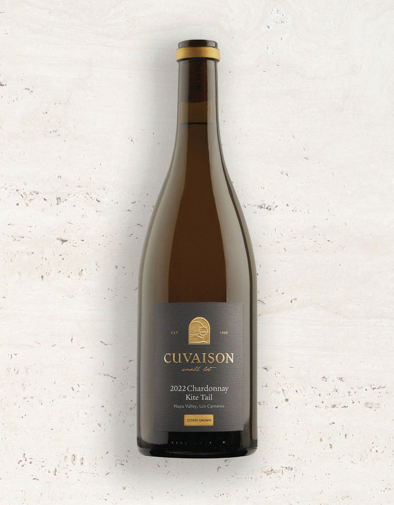 A bottle of 2022 Cuvaison Chardonnay, Kite Tail with a dark label rests on a light, textured surface. The label features the brand name, vintage year, and wine type in gold and white text.