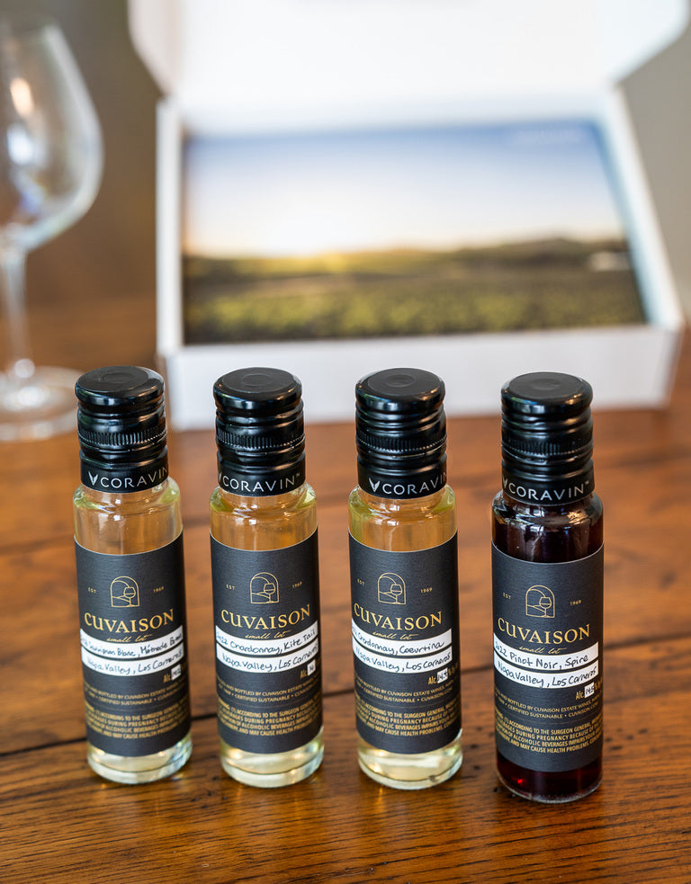Four small bottles from the Custom Tasting Kit are lined up on a wooden table. An open box with a scenic photo is behind them, and an empty wine glass sits to the left.