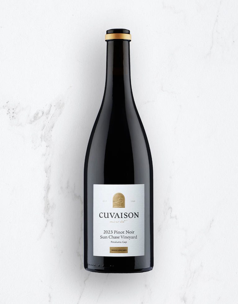 A bottle of 2023 Cuvaison Pinot Noir, Sun Chase Vineyard stands upright on a white marble surface, featuring a black foil top and a white label with gold and black text.