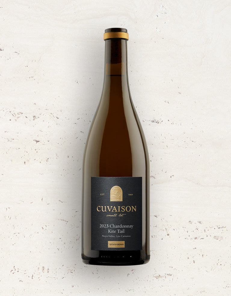 A bottle of 2023 Cuvaison Chardonnay, Kite Tail stands upright against a light textured background, featuring a dark label with gold and white text.