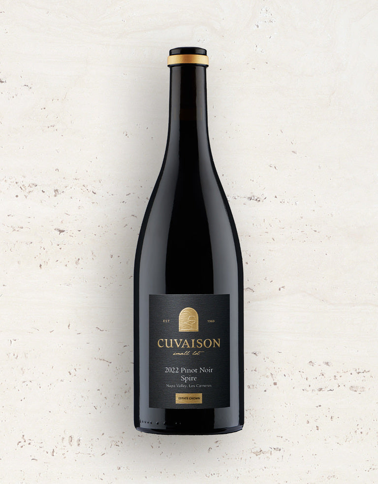 A bottle of 2022 Cuvaison Pinot Noir, Spire stands upright against a light beige textured background. Its black label features gold and white text with the wines name and brand.
