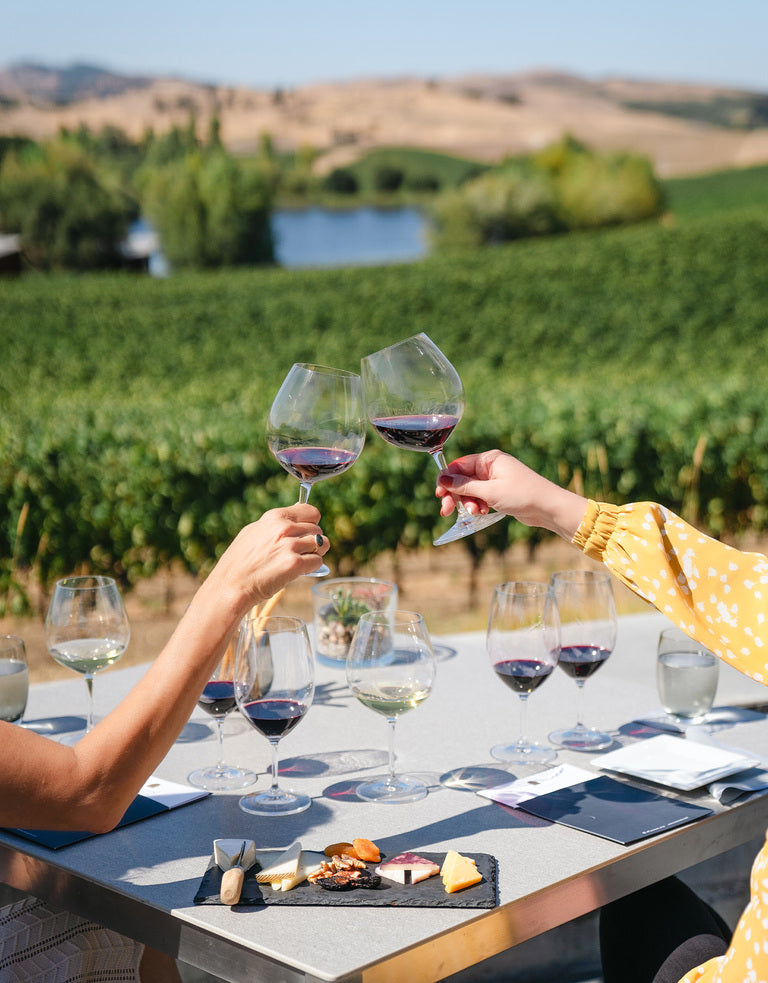 Two people enjoy the Gift the Winemakers Tasting Experience at an outdoor table with wine flights and cheese, overlooking a vineyard and rolling hills under a clear sky.