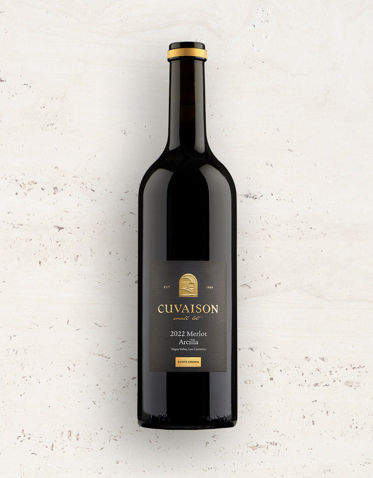 A bottle of 2022 Cuvaison Merlot, Arcilla with a black and gold label stands upright on a light textured surface.