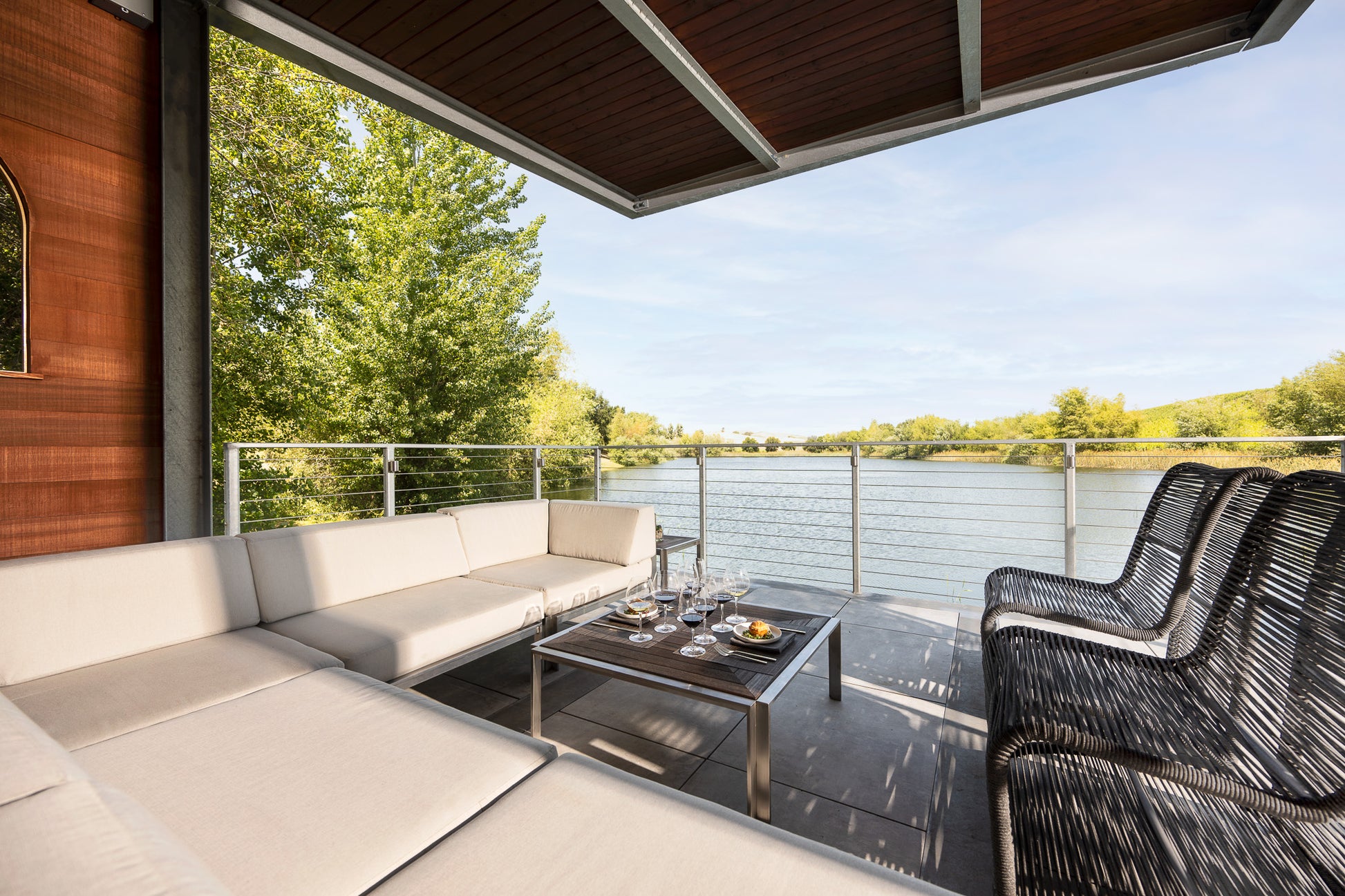 A covered outdoor patio with beige sectional seating, a black wicker chair set, and a coffee table with drinks and snacks overlooks a river and surrounding green trees under a clear sky.