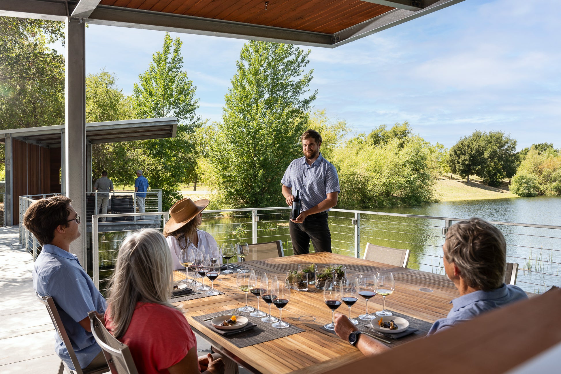 A man stands beside an outdoor dining table set with wine glasses and plates, speaking to four seated people under a covered patio near a lakeside with trees and greenery in the background.