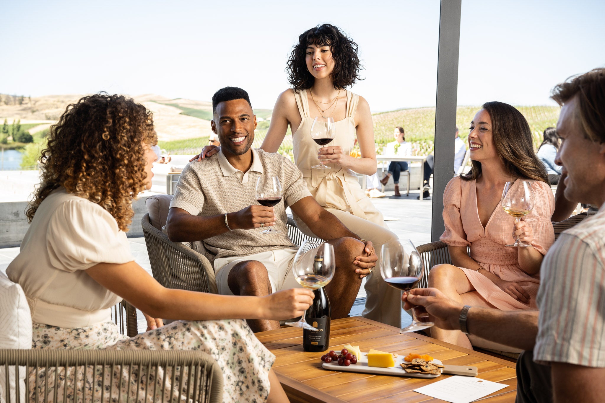 A group of five adults sit around a wooden table outdoors, holding glasses of red wine and talking. The table has a wine bottle, cheese, and fruit. Vineyards and rolling hills are visible in the background.