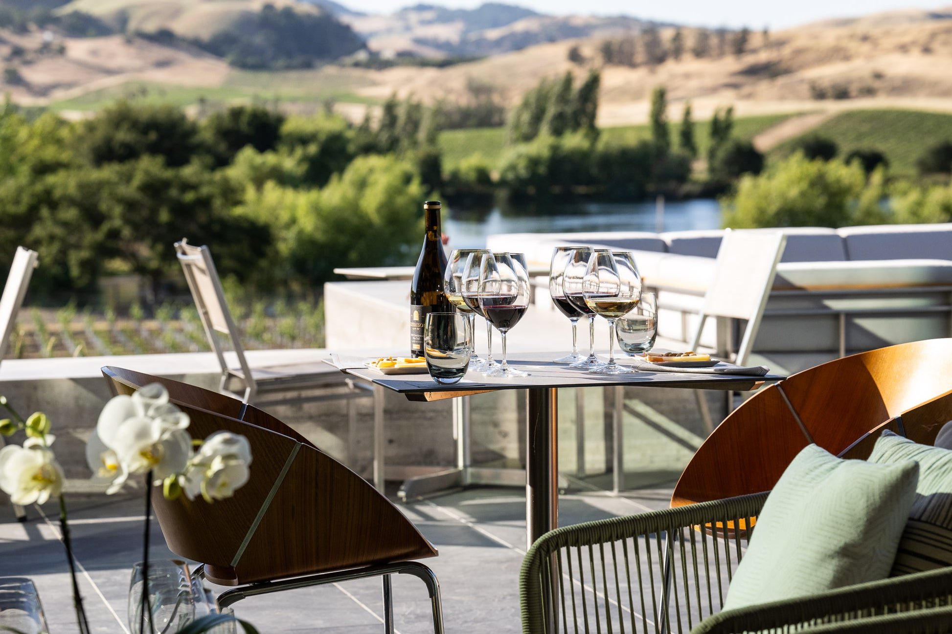 A table on a patio set with a bottle of wine, three filled wine glasses, and small plates with snacks. Modern chairs surround the table, with a scenic view of vineyards and hills in the background.