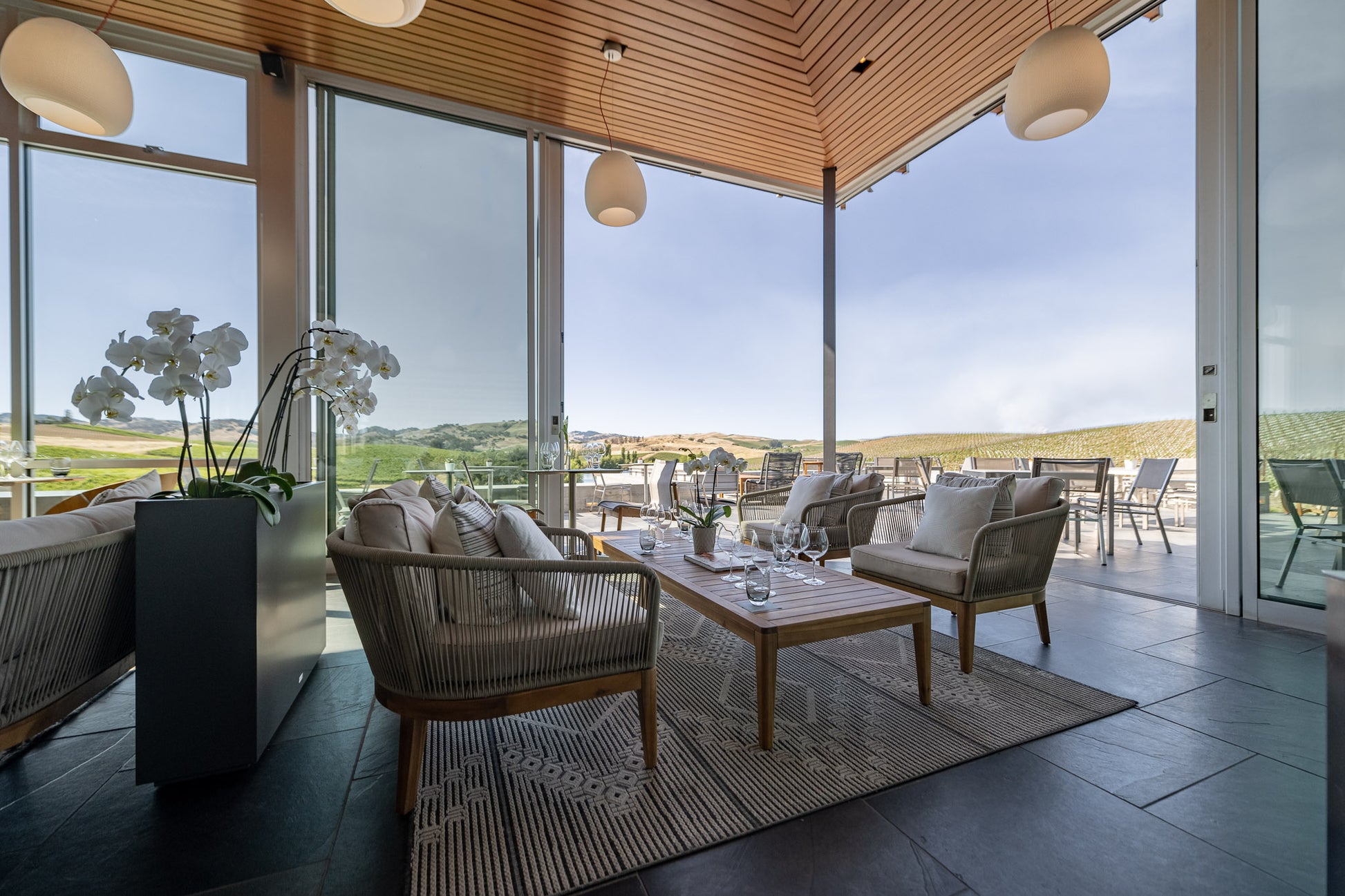 Modern lounge area with light wooden furniture, a rug, and a potted orchid, set beside floor-to-ceiling windows looking out onto a patio and green hills under a clear sky. Tables and chairs are placed outside.